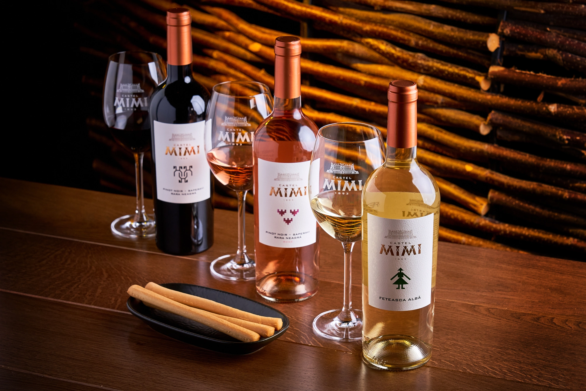 Wine bottles and glasses on a wooden table with a plate of breadsticks, against a backdrop of stacked logs.
