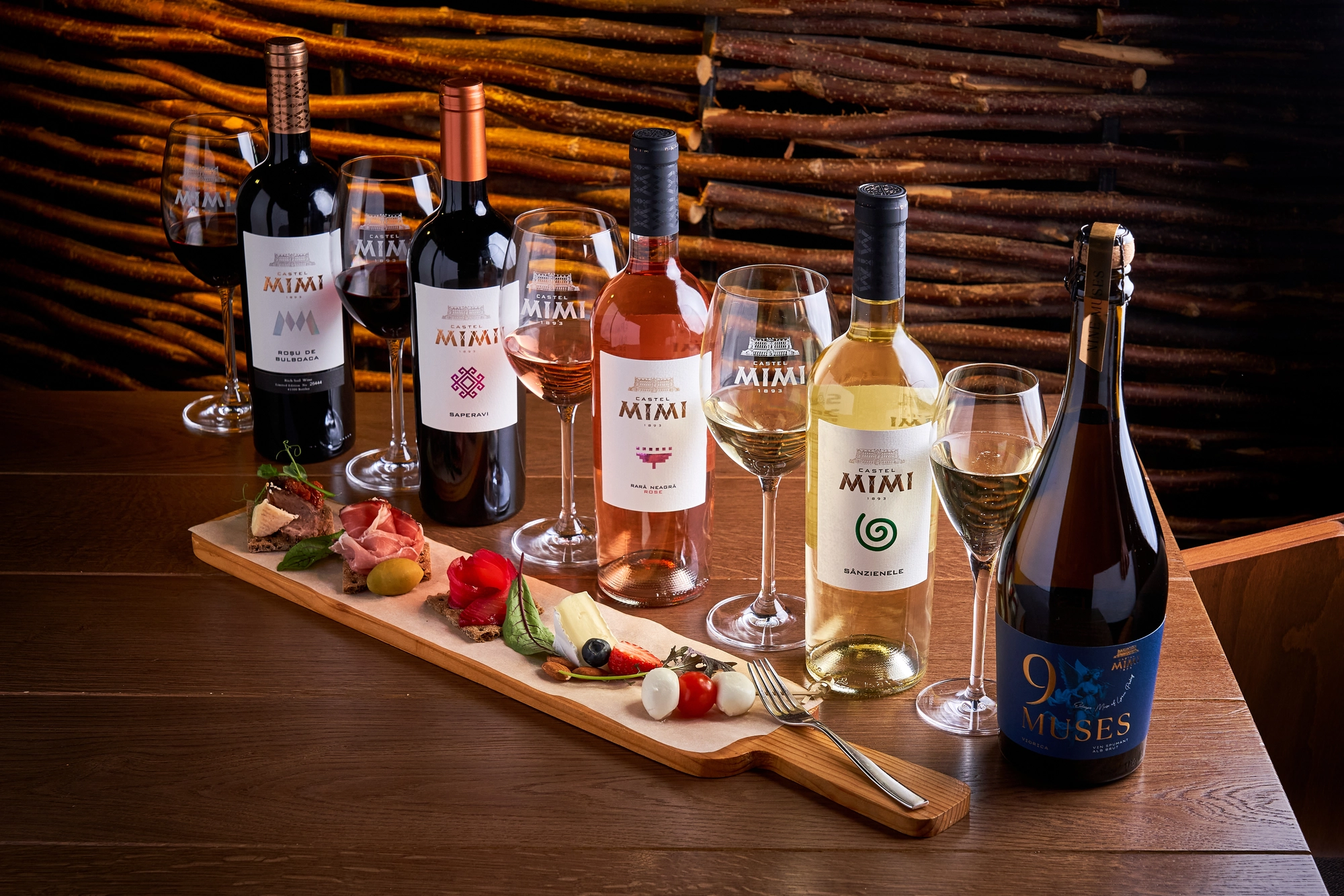 Wine bottles and stemmed glasses arranged on a wooden table beside a tasting board of bread, cheese, and vegetables, with stacked logs in the background.