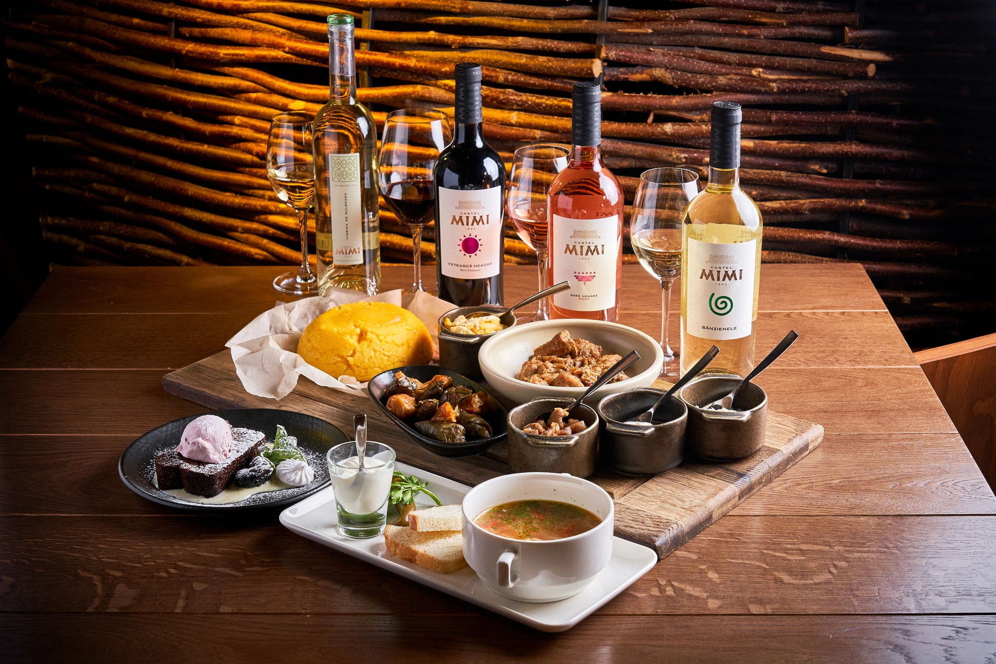 Wine bottles and glasses on a rustic wooden table with bowls of food and desserts in a warmly lit dining setting, background of stacked wood.