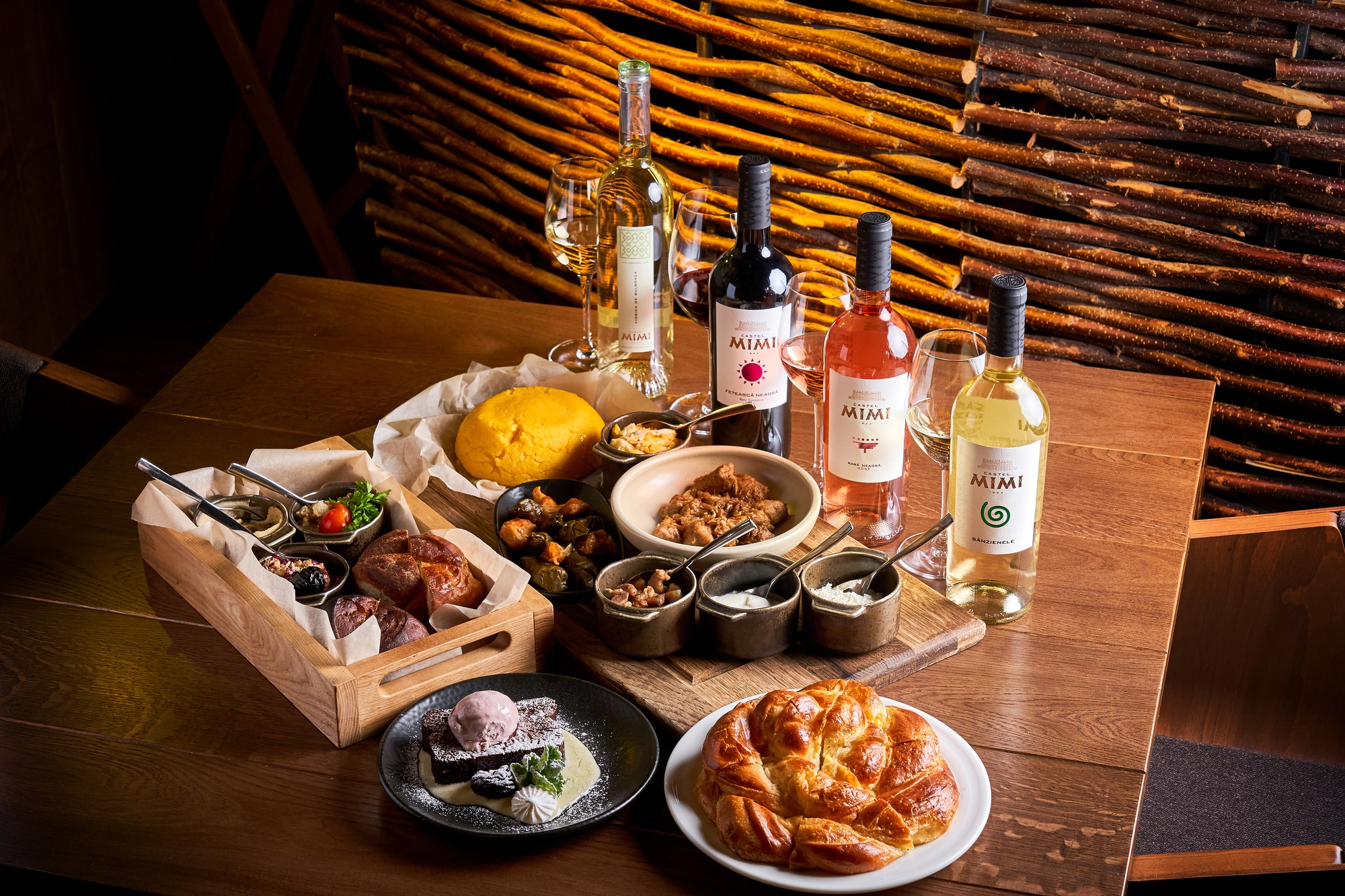 Wine bottles and glasses with a charcuterie spread on a wooden table, plus bread and desserts in a cozy setting.