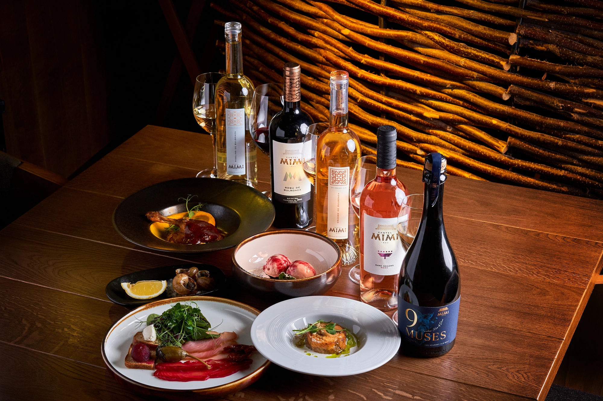 Wine bottles and glasses on a wooden table with plates of appetizers, set against a back wall of stacked wooden logs.