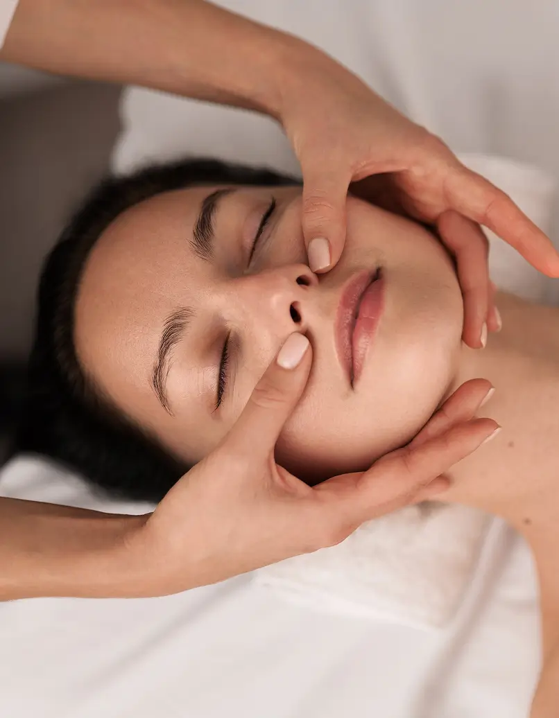 Woman lying with eyes closed while a therapist gently masses her cheeks for a facial treatment.