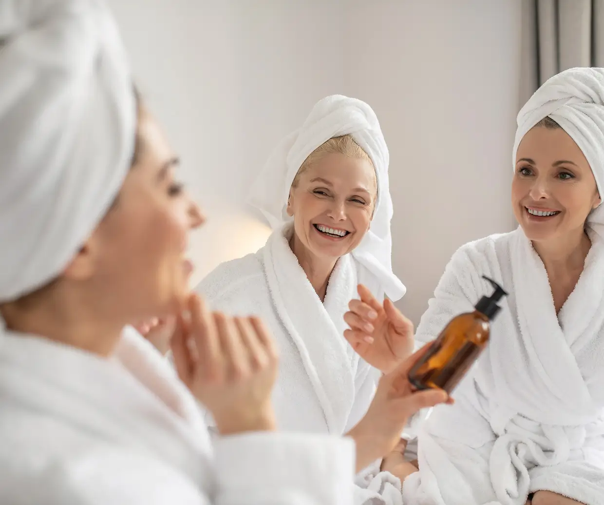 Three women in white robes and towel wraps sit together in a spa, smiling as one shows a brown pump bottle.