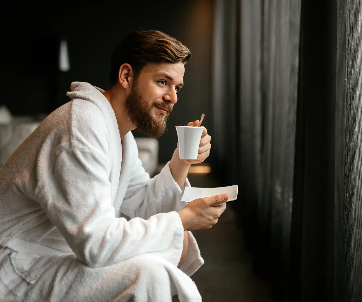 Man in a white bathrobe sipping coffee from a cup while looking out a window.