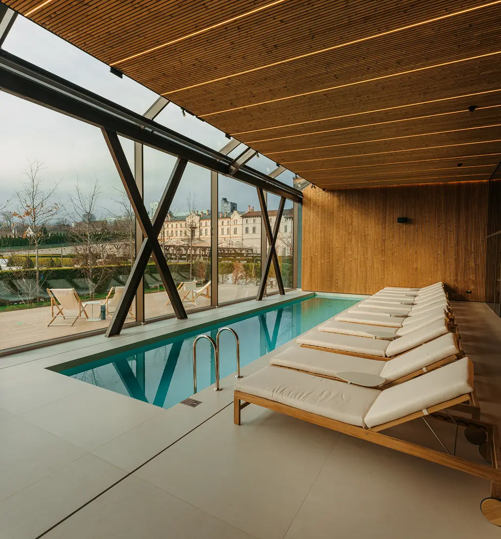 Indoor pool area with white cushioned lounge chairs along the pool, wood-paneled walls and ceiling, and large glass walls revealing an outdoor terrace.